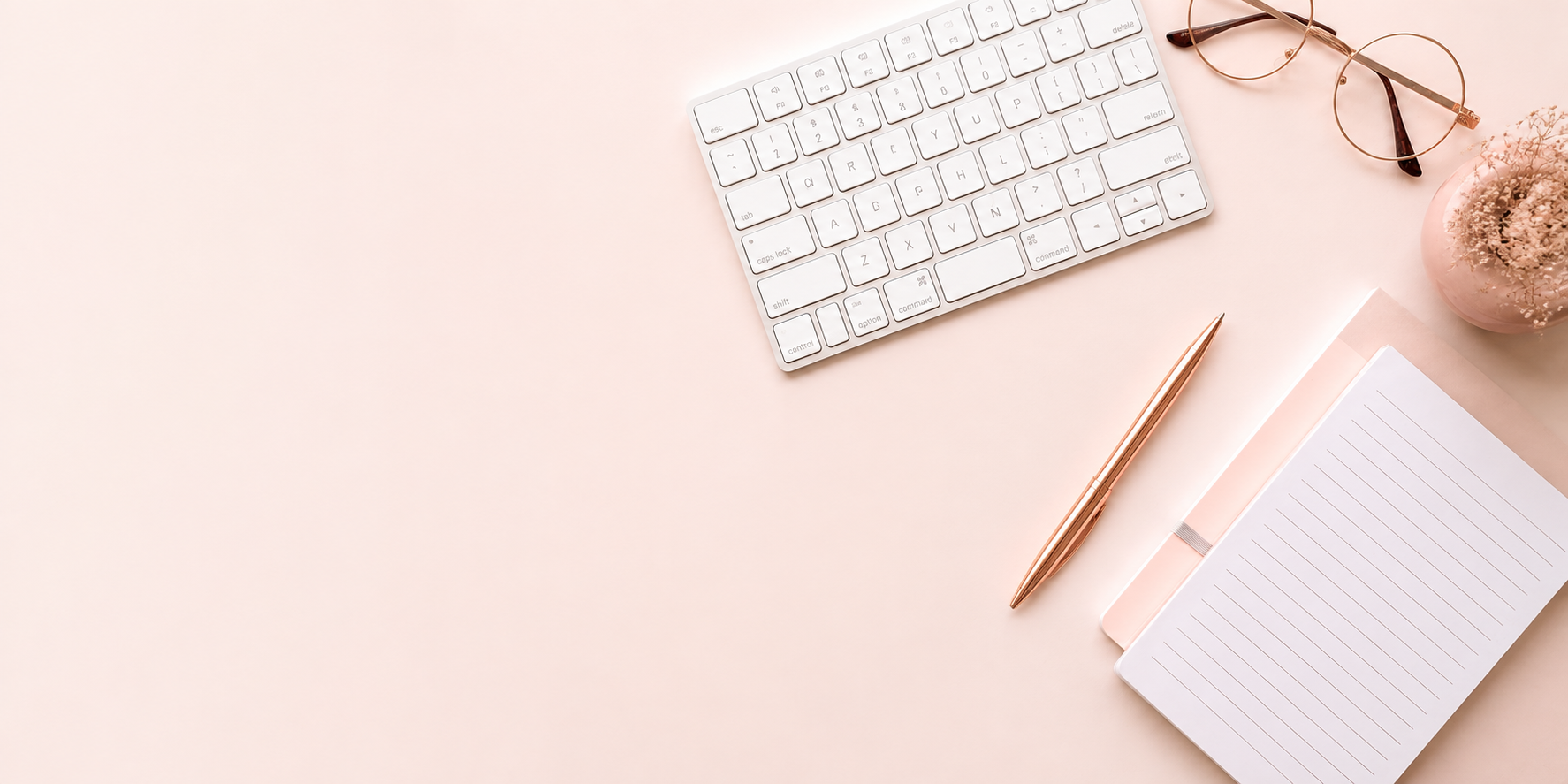 Schreibtisch von oben mit Tastatur, Notizbuch, Kugelschreiber und Brille auf rosa Untergrund